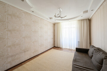Neutral room with beige patterned walls, a large window with sheer curtains, a patterned rug, and a brown sofa. A modern chandelier hangs from the white ceiling