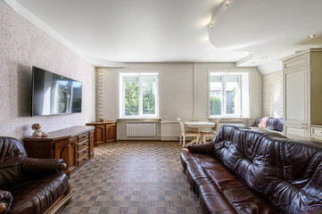 Cozy living room with brown leather furniture, TV, wooden cabinets, and two windows. A light-colored kitchen area is visible in the background