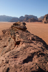A panoramic landscape shot of a vast, rocky desert with a prominent natural arch and sculpted rock formations in the foreground, set against a clear blue sky. This image captures the monumental scale 