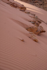 A low-angle close-up of a pinkish-orange sand dune with a sharp, ridged surface from wind erosion, leading up to a protruding formation of jagged, rusty-red rocks. This image captures the powerful con