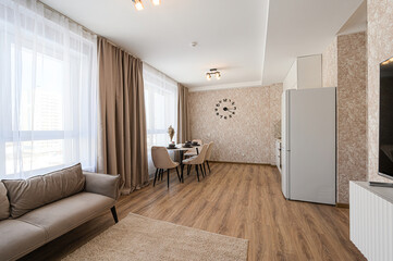 bright, modern living space with a beige sofa, dining set, large clock, and kitchen appliances