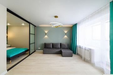 Bright, modern apartment with a gray sofa, light wood floors, teal accents, and a glass-enclosed bedroom. Stylish lighting and a clean design enhance the space