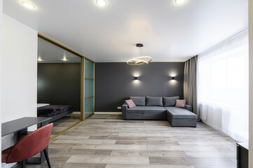 Modern apartment interior featuring a gray sofa, wood floors, and a bedroom view through a sliding glass door. Contemporary lighting fixtures add style