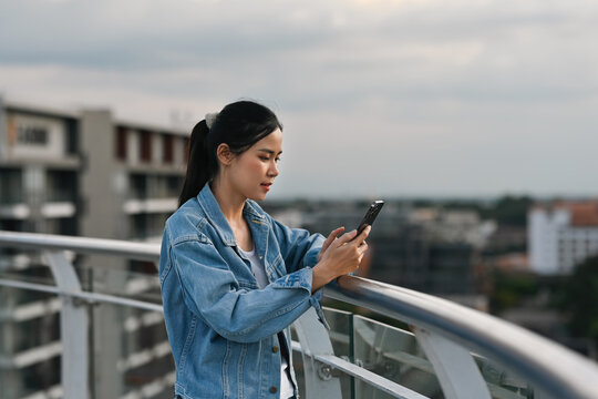 A Young Asian woman in a denim jacket smiles as she browses her smartphone, enjoying a moment outdoors on a city balcony