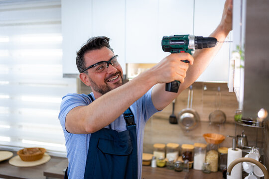Man installs cabinet in modern kitchen using electric drill while wearing safety glasses