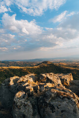 A rocky mountain top with a cloudy sky in the background. The sky is blue with some clouds scattered throughout. The mountain is covered in trees and the rocks are large and jagged