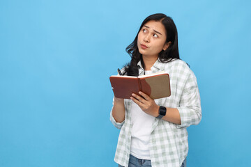 Young Asian woman wearing casual shirt, writing in notebook, remembering something looking up in empty space, and making dream list isolated on blue background.