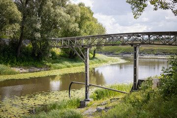 Fototapeta premium Metal frame structure over a calm river surrounded by lush green vegetation during a sunny day in early summer