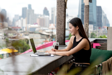 Fototapeta premium Asian young adult woman working on laptop at city rooftop cafe productive professional remote work lifestyle digital nomad urban setting