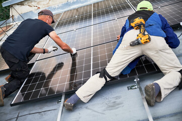 Workers building solar panel system on metal rooftop of house. Two men installers installing photovoltaic solar module outdoors. Alternative, green and renewable energy generation concept.