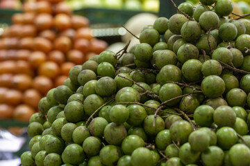 Close-up of green mamoncillo fruits stacked at a market stall, with tamarillos blurred in the background.