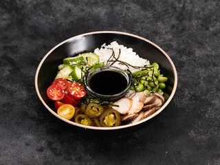 A colorful rice bowl with fresh vegetables: tomatoes cherry, cucumbers, beans, chicken meat, aromatic herbs, and a savory dipping sauce for enhanced flavor on black background.