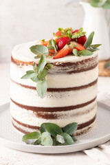 Fresh strawberries and eucalyptus sprigs adorn a delicious, rustic naked chocolate cake with creamy white frosting on a gray plate.