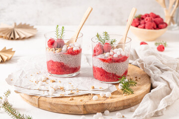 Two glasses of layered raspberry and chia seed pudding topped with fresh raspberries and white crowberry on a rustic wooden platter