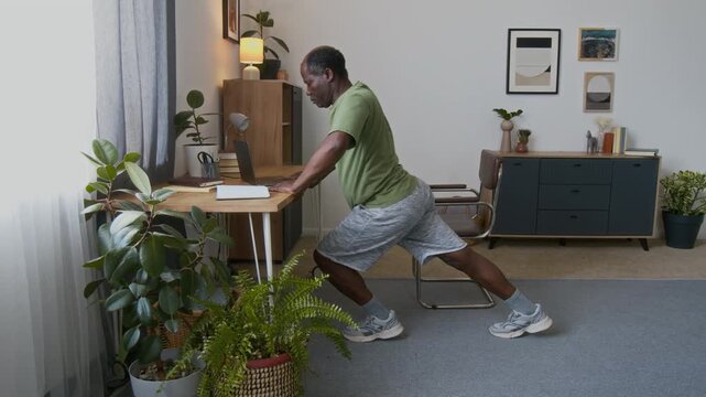 Full side shot of middle-aed African American man incasual clothes leaning on esk, doing press-ups and font leg lunge movements totrain sore body muscles, wile working on computer