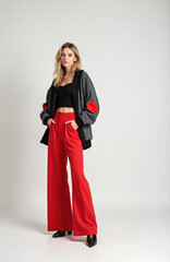 Blonde woman wearing black crop top, grey jacket with red accents, and red flared pants. Posing confidently on white studio background. Fashionable businesswoman, stylish modern look.