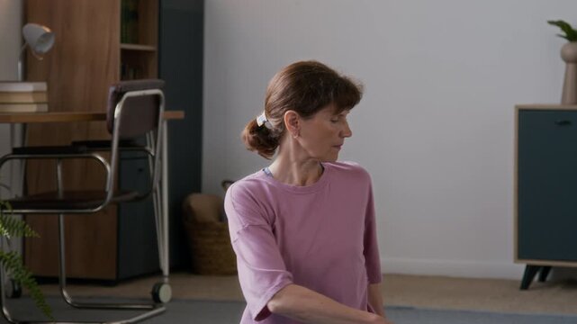 Medium shot of senior Caucasian woman in pink t-shirt doing spine rotation and torso twist exercise while sitting on floor in living room, warming up for daily home fitness workout