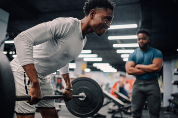 Determined male athlete lifting weights while personal trainer supervises in gym