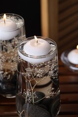 Burning candles, spa stones and flowers in glasses of water on wooden table against black background, closeup
