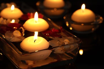 Burning candles and orchid flowers in glass container of water against black background, closeup