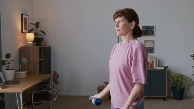 Medium close-up of senior Caucasian woman in pink t-shirt doing repeats of bicep curl exercise using dumbbells in living room, paying attention to safety technique during home workout