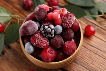 Mix of ripe frozen berries in bowl and green leaves on wooden table, closeup