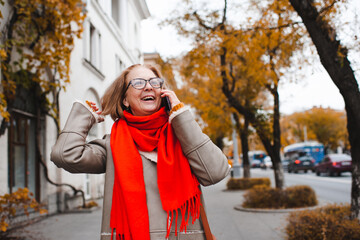 Smiling mature woman 55-60 year old wearing eye glasses, knitted red scarf and jacket in city street outdoors. Autumn season. Senior lifestyle.