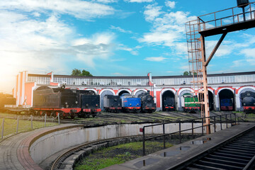 Naklejka premium Many old steam locomotives are parked inside a vintage red and white roundhouse in Eastern Europe. Tracks and maintenance equipment surround the trains