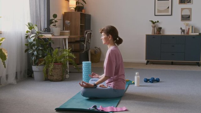 Full side shot of sporty senior woman in pink t-shirt and blue leggings sitting on floor in lotus posture, hands in mudra, meditating and doing pranayama breathing practice in living room at home