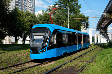 Naklejka premium A bright blue tram moves along its tracks in an urban setting on a sunny day. Apartment buildings and an elevated structure are visible in the background
