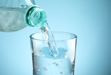 Pouring refreshing soda water from bottle into glass at table against light blue gradient background, closeup