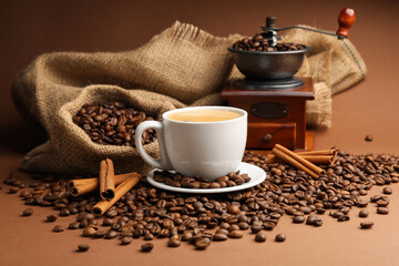 Cup of hot coffee, beans, grinder and cinnamon on brown background