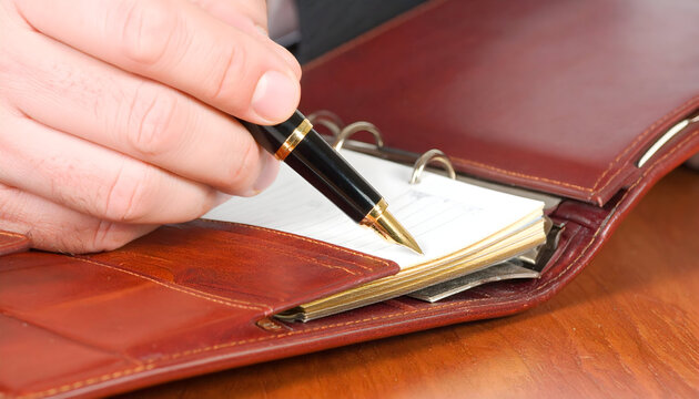 Close-up of a hand writing with a pen in a leather personal organizer on a wooden desk