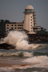 A powerful wave crashes onto the sandy shore, reaching the exterior of a hotel in Sri Lanka during sunset.