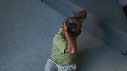 Medium full shot of sporty elderly African American man in t-shirt, shorts and trainers performing repeats of abdominal crunches with torso rotation on floor at home during daily workout - Powered by Adobe