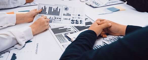 A diverse group of professionals engage in a collaborative business meeting, analyzing charts and data on a table. Their hands indicate teamwork and strategic discussion. SACTR