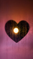 A heart shape is drawn on a rain streaked window, with a bright light seen through it