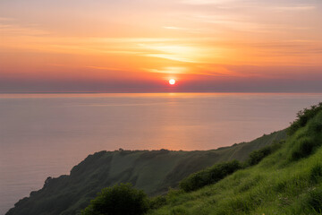 A vibrant sunset over the ocean, with a green, grassy cliff visible on the right side of the frame