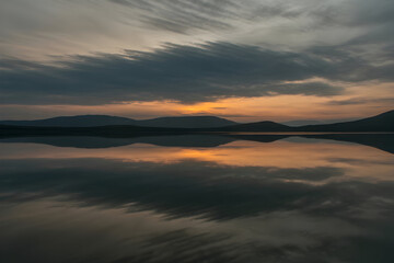 Still water perfectly mirrors a dramatic, cloud streaked sunset over distant mountains, emphasizing natures reflective beauty