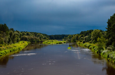 A calm river flowing through lush green forest under dramatic storm clouds and sunlight.