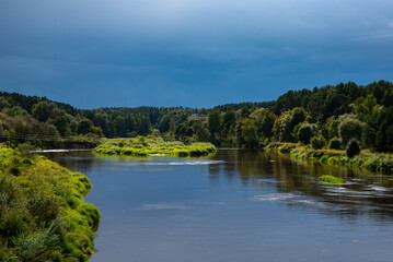 A calm river winding through lush green forests and meadows under a dramatic blue stormy sky.