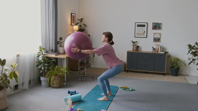 Wide shot of elderly Caucasian woman in pink t-shirt, blue leggings performing squats while holding out balance ball in living room at home during daily fitness routine to maintain health in old age