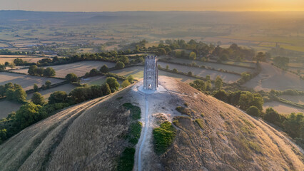 An aerial drone shot of Glastonbury Tor with St Michael’s Tower lit by the rising sun. Rolling farmland and trees surround the historic hilltop, creating a serene and atmospheric scene. © Jez