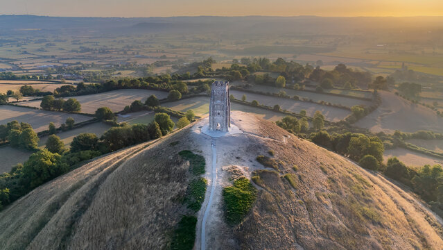 An aerial drone shot of Glastonbury Tor with St Michael&rsquo;s Tower lit by the rising sun. Rolling farmland and trees surround the historic hilltop, creating a serene and atmospheric scene.