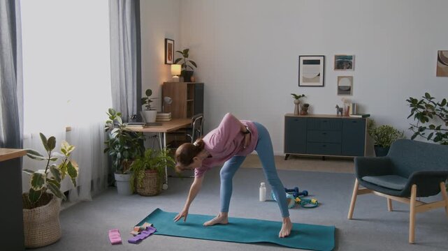 Full shot of sporty elderly Caucasian woman in pink tshirt and blue leggings practicing tai chi movements while standing on fitness mat in living room, during daily home workout in retirement