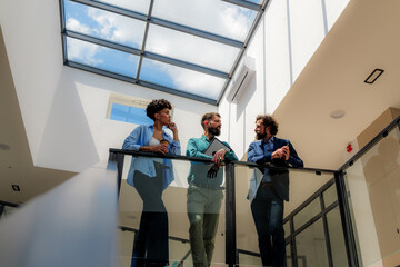 Three business people are enjoying a conversation on the balcony of their modern office building, illuminated by natural light from the skylight above