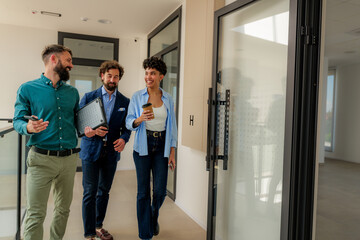 Three cheerful businesspeople are walking in the office hallway, holding laptop, smartphone and takeaway coffee, discussing business and smiling