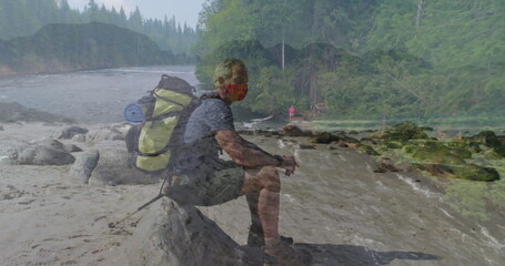 Sitting hiker wearing red mask on fallen log by flowing river, carrying backpack with rolled pad
