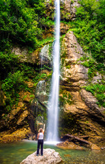 Unknown blonde woman with her back turned, pointing to an 18-meter waterfall that cascades into a small lagoon.