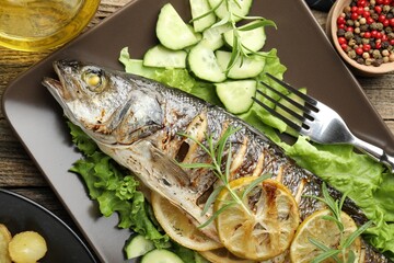 Tasty roasted seabass fish served with vegetables and lemon on wooden table, flat lay. Homemade seafood dish